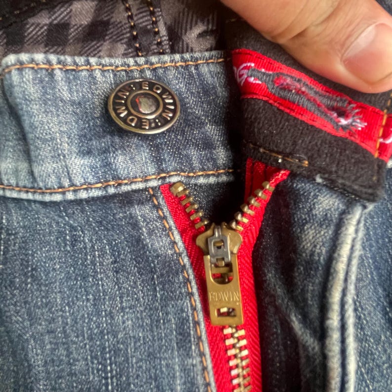 K&ouml;nnte beinhalten: Nahaufnahme einer blauen Jeans mit rotem Rei&szlig;verschluss und Metallknopf. Der goldfarbene Rei&szlig;verschluss tr&auml;gt den Markennamen "EDWIN". Der Knopf tr&auml;gt ebenfalls den Markennamen "EDWIN". Die Jeans hat innen ein Karomuster.