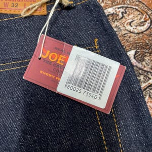May include: Close-up of dark blue denim jeans with a tag. The tag is red with the word "JOE" in large yellow letters. The tag also has the text "THE CAT" and a barcode. The jeans are size W32.