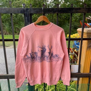 May include: A coral-colored sweatshirt with a crew neck and long sleeves. The front features a detailed illustration of a herd of elk in a dark blue hue. The sweatshirt is hanging on a wooden hanger.