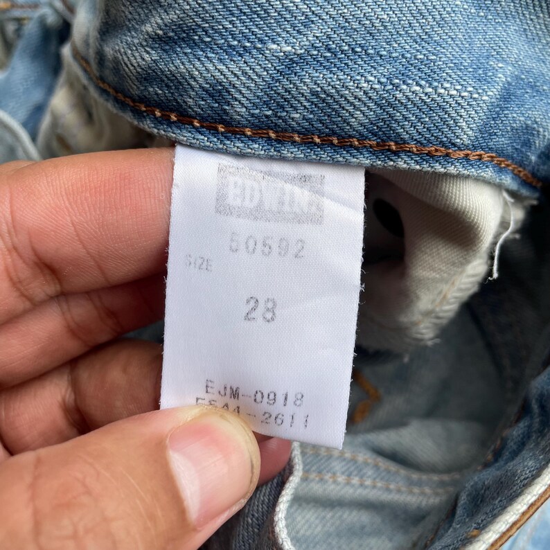 May include: Close-up of a light blue denim jeans tag. The tag displays the number "28" indicating the waist size. Other visible text includes "50592", "EJM-0918", and "F544-2611". The jeans are vintage.
