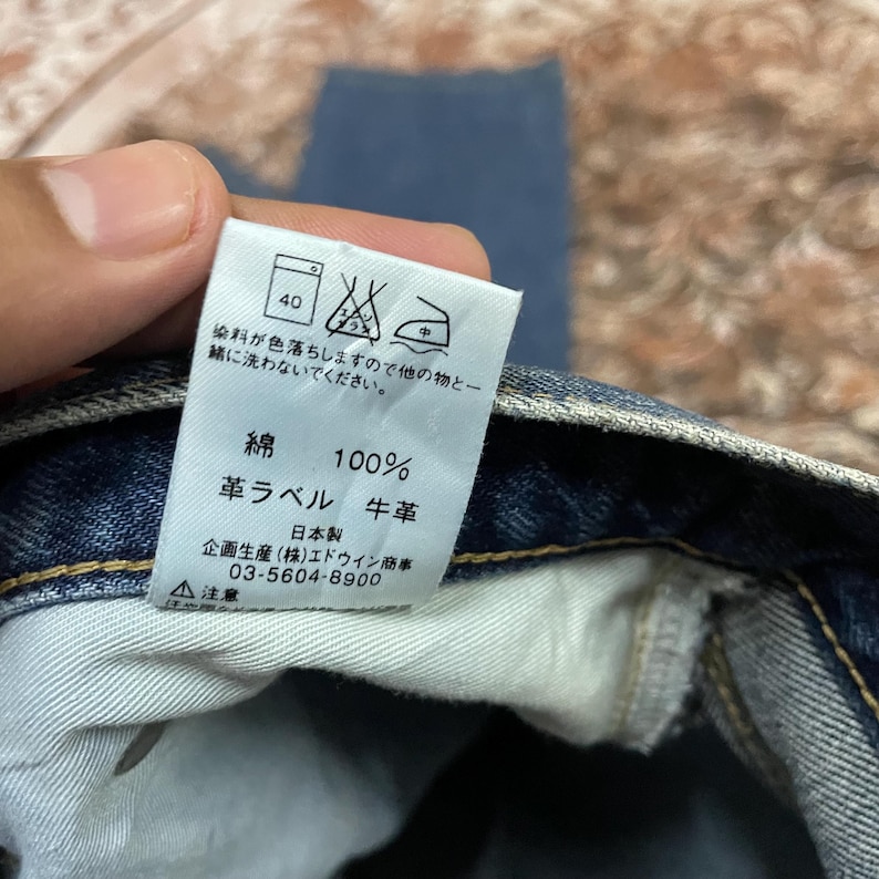 Puede incluir: Primer plano de una etiqueta blanca con texto japon&eacute;s y s&iacute;mbolos de cuidado, adherida a una tela vaquera azul. La etiqueta indica que la tela es 100% algod&oacute;n con una etiqueta de cuero. Tambi&eacute;n se incluyen instrucciones de lavado.