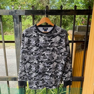 Könnte beinhalten: Ein langärmeliges Camouflage-Muster-Shirt in Grau- und Schwarztönen mit Rundhalsausschnitt und schwarzen Bündchen. Das Shirt hängt an einem Holzbügel. Das Polo-Logo ist auf der Brust zu sehen.