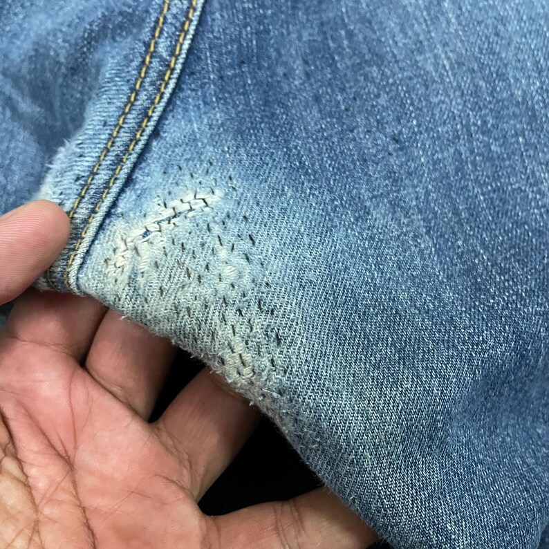 May include: A pair of blue denim jeans featuring a visible repair area with dense, dark stitching over a faded, worn section of the fabric. The repair is located near a seam with gold-colored thread, showcasing a textured, reinforced mend.