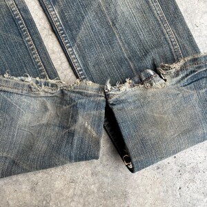 May include: Close-up of blue denim jeans with distressed, frayed hems. The jeans have a faded wash and show signs of wear, with visible stitching and a folded cuff. The fabric appears worn and textured.