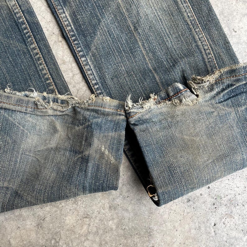 May include: Close-up of blue denim jeans with distressed, frayed hems. The jeans have a faded wash and show signs of wear, with visible stitching and a folded cuff. The fabric appears worn and textured.