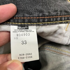 May include: Close-up of a tag from Wrangler jeans. The tag is white with black text, displaying the brand name, style number W04903, size 33, and other details. The jeans are dark gray.