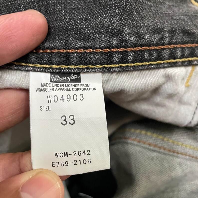 May include: Close-up of a tag from Wrangler jeans. The tag is white with black text, displaying the brand name, style number W04903, size 33, and other details. The jeans are dark gray.