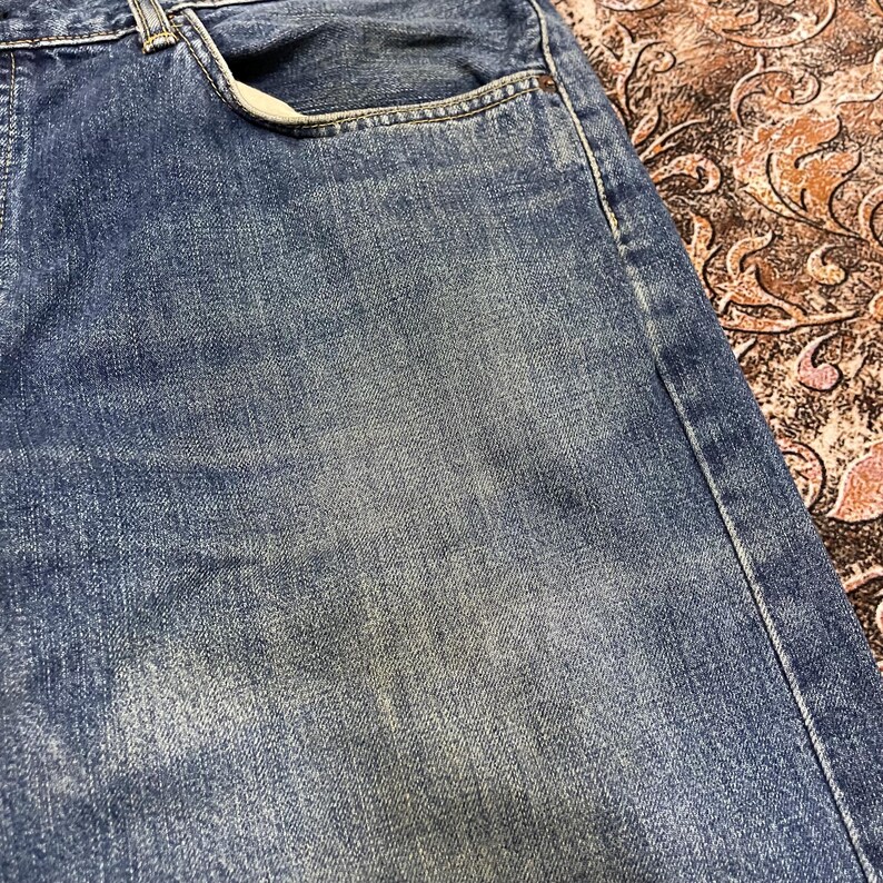 May include: A pair of blue denim jeans featuring a classic five-pocket construction with a faded wash effect. The fabric displays a textured weave with visible stitching along the seams and a standard waistband with a front pocket opening.