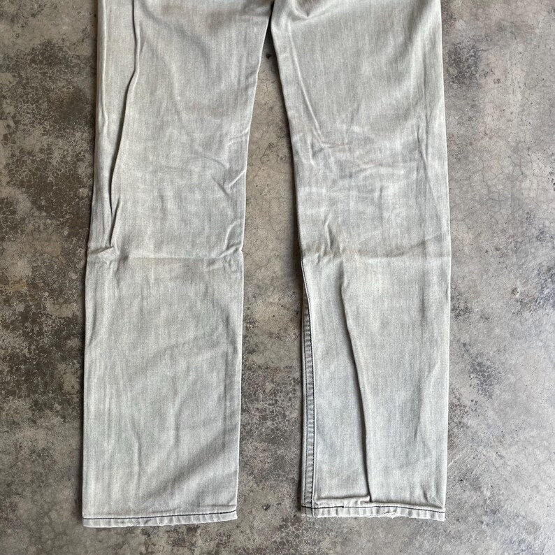 K&ouml;nnte beinhalten: Hellgraue Jeans aus Denim. Die Jeans hat einen geraden Beinverlauf und ein verwaschenes Aussehen. Der Stoff weist eine subtile Textur und Abnutzung auf. Die Jeans liegt flach auf einer neutralen Oberfl&auml;che.