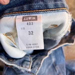 Pu&ograve; includere: Primo piano dell'interno di un paio di jeans blu. Un'etichetta bianca mostra il marchio "EDWIN", il numero di modello "403" e la taglia "32". Sono visibili anche i codici "EIN-4301" e "E566-2709".