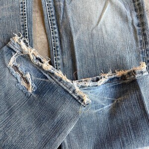 May include: Close-up of light blue denim jeans with distressed, frayed hems. The jeans show signs of wear and tear, with visible fading and a textured appearance. The fabric is a classic denim weave.