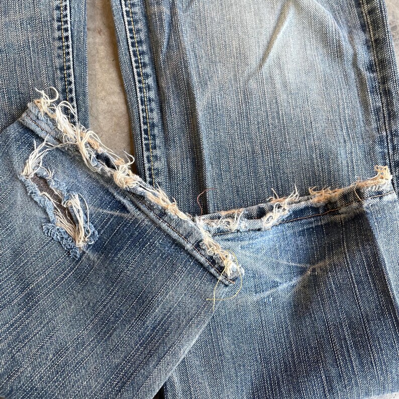 May include: Close-up of light blue denim jeans with distressed, frayed hems. The jeans show signs of wear and tear, with visible fading and a textured appearance. The fabric is a classic denim weave.