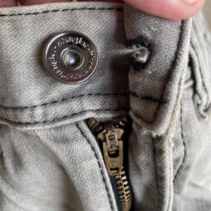 K&ouml;nnte beinhalten: Nahaufnahme eines hellgrauen Denim-Stoffs mit einem silbernen Knopf und einem Messingrei&szlig;verschluss. Der Knopf ist mit dem Wort "Wrangler" gepr&auml;gt. Der Rei&szlig;verschluss ist teilweise ge&ouml;ffnet und zeigt die Messingz&auml;hne.