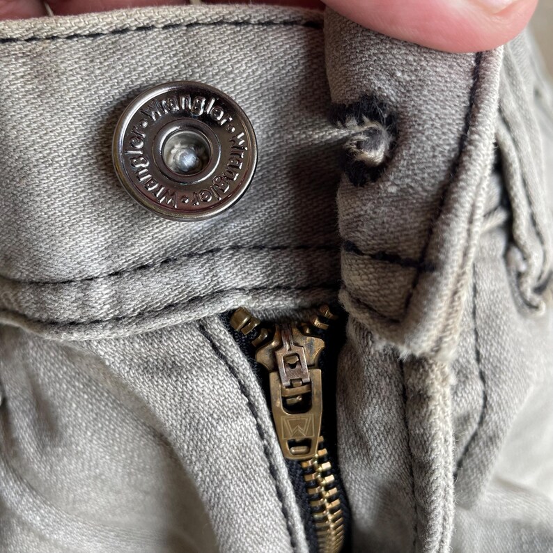 K&ouml;nnte beinhalten: Nahaufnahme eines hellgrauen Denim-Stoffs mit einem silbernen Knopf und einem Messingrei&szlig;verschluss. Der Knopf ist mit dem Wort "Wrangler" gepr&auml;gt. Der Rei&szlig;verschluss ist teilweise ge&ouml;ffnet und zeigt die Messingz&auml;hne.