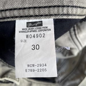 K&ouml;nnte beinhalten: Nahaufnahme eines Wrangler Jeans-Etiketts. Das wei&szlig;e Etikett hat schwarzen Text mit dem Wrangler-Logo, Produktinformationen und der Gr&ouml;&szlig;e 30. Die Jeans hat eine hellgraue Farbe.