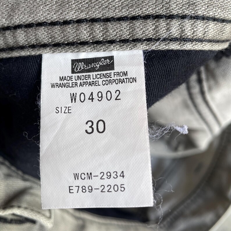 K&ouml;nnte beinhalten: Nahaufnahme eines Wrangler Jeans-Etiketts. Das wei&szlig;e Etikett hat schwarzen Text mit dem Wrangler-Logo, Produktinformationen und der Gr&ouml;&szlig;e 30. Die Jeans hat eine hellgraue Farbe.