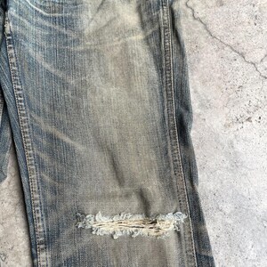 May include: Close-up of distressed blue denim jeans with a large, frayed hole at the knee. The jeans have a faded, worn appearance with visible stitching and subtle color variations, suggesting a vintage or well-loved aesthetic.