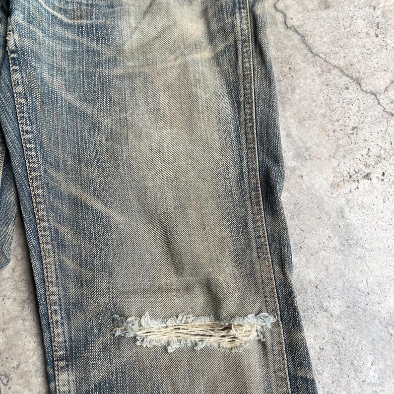 May include: Close-up of distressed blue denim jeans with a large, frayed hole at the knee. The jeans have a faded, worn appearance with visible stitching and subtle color variations, suggesting a vintage or well-loved aesthetic.