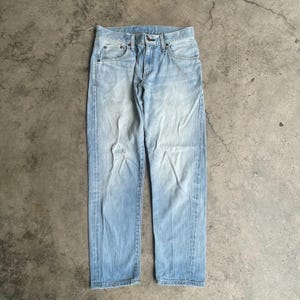 May include: Faded light blue denim jeans with a distressed appearance. The jeans feature a classic five-pocket design, a button and zip closure, and a small tear on the left leg. The jeans are displayed flat on a concrete surface.