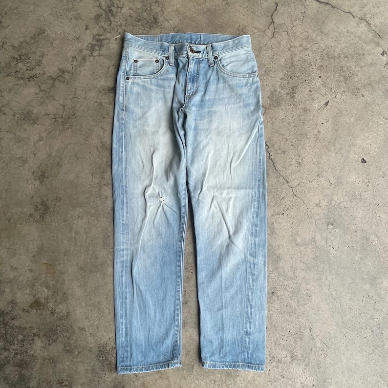 May include: Faded light blue denim jeans with a distressed appearance. The jeans feature a classic five-pocket design, a button and zip closure, and a small tear on the left leg. The jeans are displayed flat on a concrete surface.