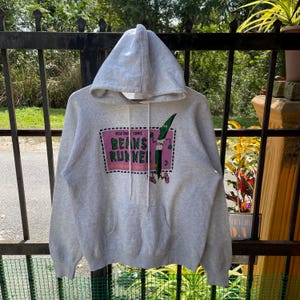 Op de afbeelding: Een grijs hoodie met een afbeelding van een groene boon die rent. De afbeelding is in een roze en groene rechthoek met de tekst "VEGETABLE COMIC BEANS RUNNER" in zwart.