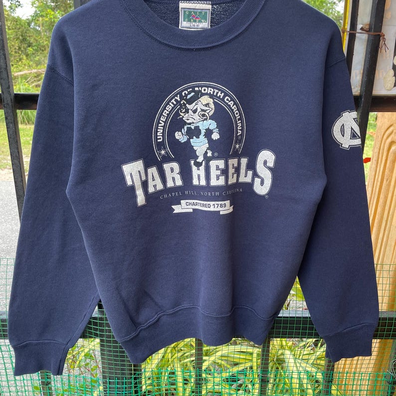 May include: Navy blue sweatshirt featuring the University of North Carolina Tar Heels logo. The logo includes a cartoon ram and the text "Tar Heels" in white. The sweatshirt also has a small logo on the sleeve.