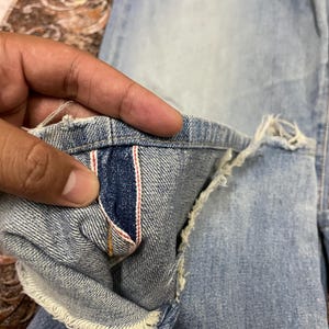 May include: Close-up of light blue denim jeans with a distressed hem. The interior pocket lining features a dark blue fabric with red and white stripes. The jeans show signs of wear and tear.