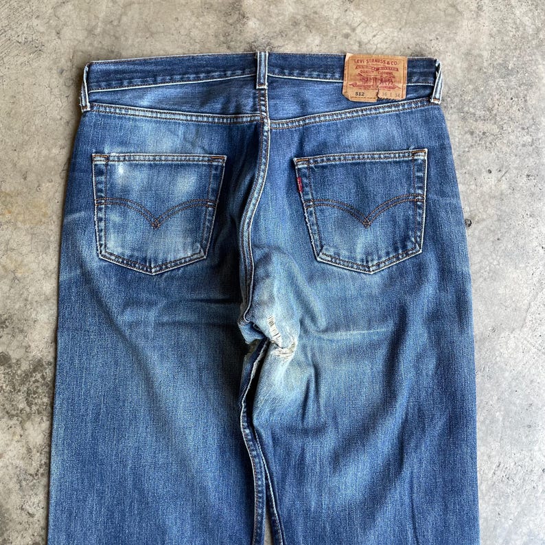 May include: Rear view of blue denim jeans featuring two back pockets and a red Levi Strauss & Co. label. The jeans have a faded look and a classic five-pocket design. The label reads "512".