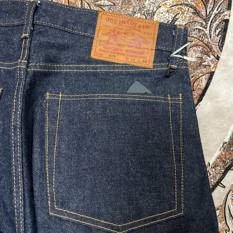 May include: Close-up of dark blue denim jeans with yellow stitching. A brown leather patch on the back pocket reads "JOE McCoy & co." and "Every Garment Guaranteed." The jeans are labeled Lot 906, W32 L36.