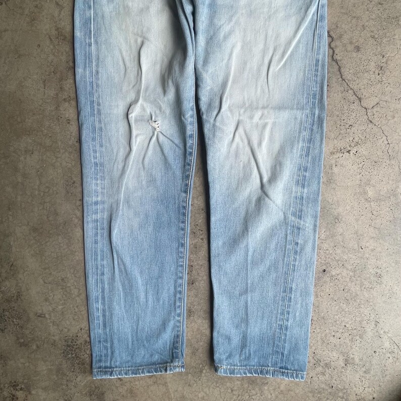 May include: Pale blue denim jeans with a faded wash and distressed details. The jeans have a classic five-pocket design and a straight leg fit. The fabric shows signs of wear and tear, adding to the vintage aesthetic.