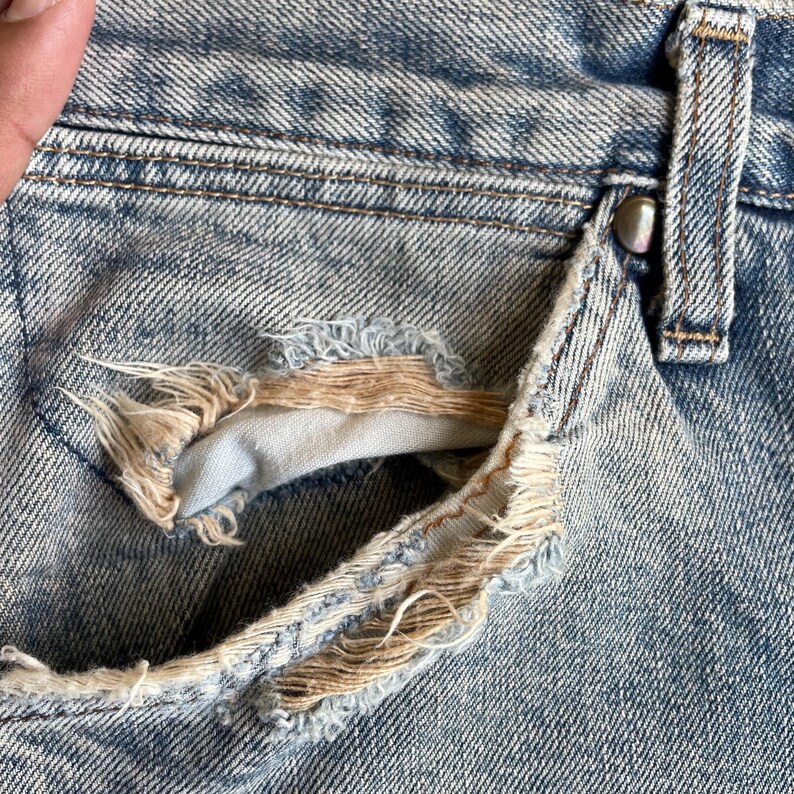 May include: Close-up of distressed light blue denim jeans. The pocket has a large tear with frayed edges, revealing a lighter fabric underneath. The jeans show signs of wear and tear.