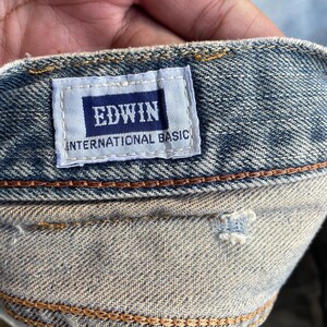 May include: Close-up of denim fabric with a rectangular label. The label, with a navy blue background, displays the brand name "EDWIN" in white capital letters, and "INTERNATIONAL BASIC" below. The denim is a light blue wash with visible stitching.