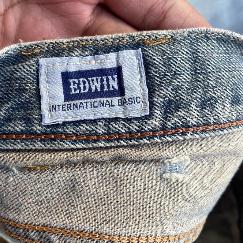 May include: Close-up of denim fabric with a rectangular label. The label, with a navy blue background, displays the brand name "EDWIN" in white capital letters, and "INTERNATIONAL BASIC" below. The denim is a light blue wash with visible stitching.
