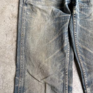 May include: Close-up of light blue denim jeans with faded areas and visible wear. The jeans show signs of use, with lighter patches and subtle texture variations. The seams are visible, and the overall look is vintage.
