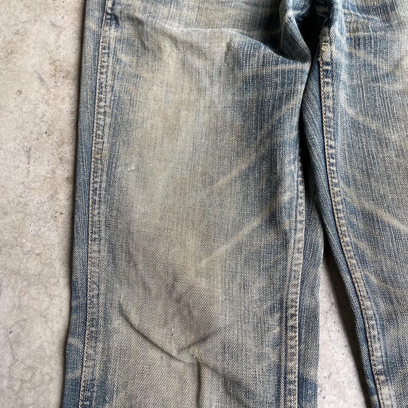 May include: Close-up of light blue denim jeans with faded areas and visible wear. The jeans show signs of use, with lighter patches and subtle texture variations. The seams are visible, and the overall look is vintage.