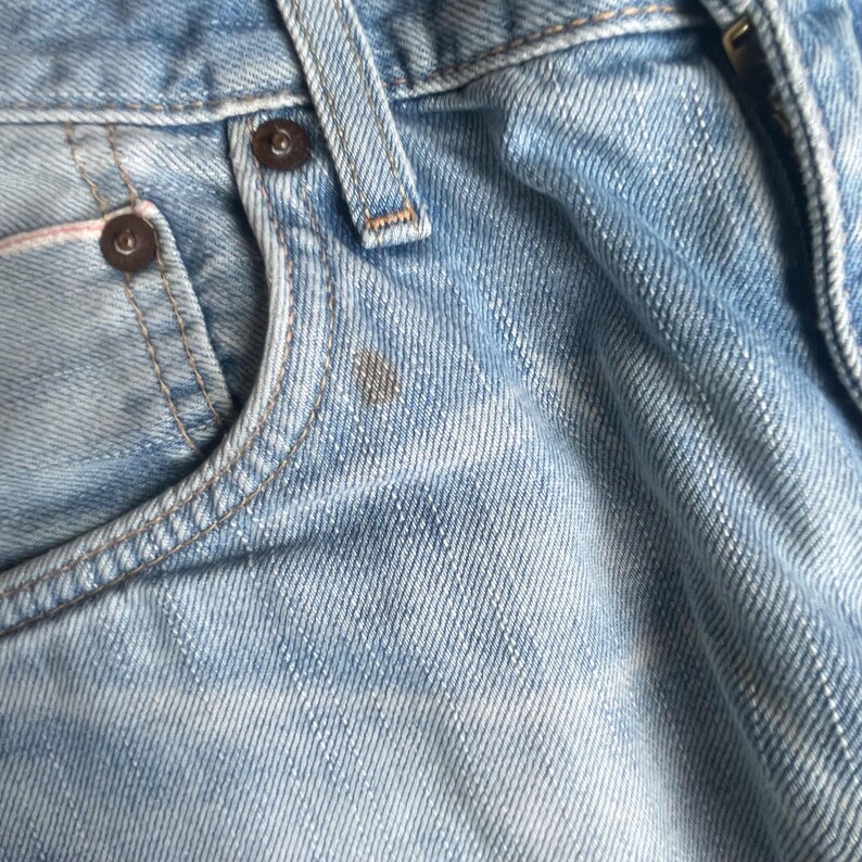 May include: Close-up of light blue denim jeans. The image shows the front pocket, belt loop, and button details. The denim has a faded, worn appearance with a small stain near the pocket.