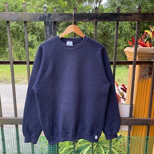 Könnte beinhalten: Ein marineblaues Sweatshirt mit Rundhalsausschnitt hängt an einem Holzbügel. Das Sweatshirt hat lange Ärmel und ein kleines weißes Etikett unten rechts. Der Hintergrund umfasst einen Metallzaun und Grün.