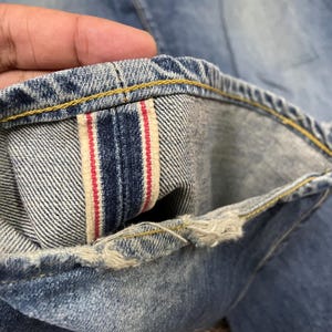 May include: A pair of light blue denim jeans featuring a visible red-line selvedge ID on the interior cuff. The hem shows signs of fraying and wear, with contrasting yellow stitching along the edge of the denim fabric and the selvedge seam.