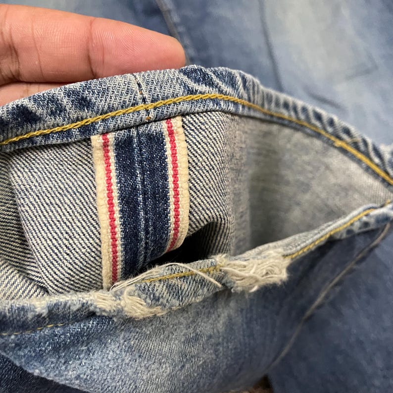 May include: A pair of light blue denim jeans featuring a visible red-line selvedge ID on the interior cuff. The hem shows signs of fraying and wear, with contrasting yellow stitching along the edge of the denim fabric and the selvedge seam.