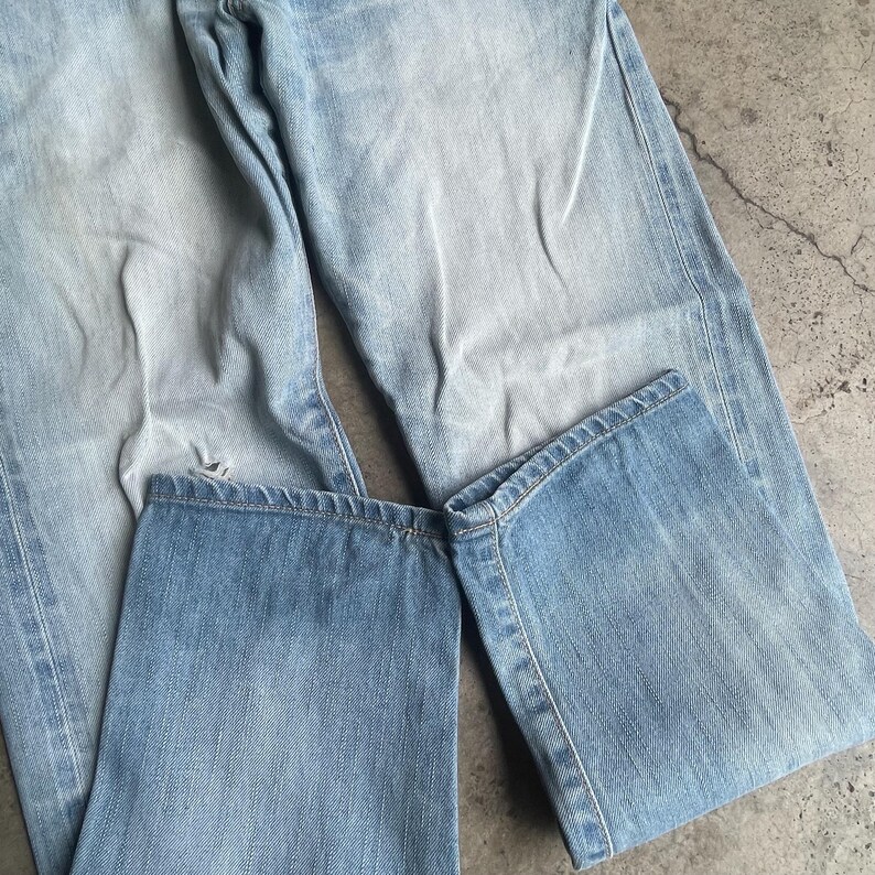 May include: Light blue denim jeans, folded and laid flat. The jeans have a faded wash and visible stitching details. The fabric appears worn, with a slight distressed look. The jeans are a classic style, suitable for casual wear.