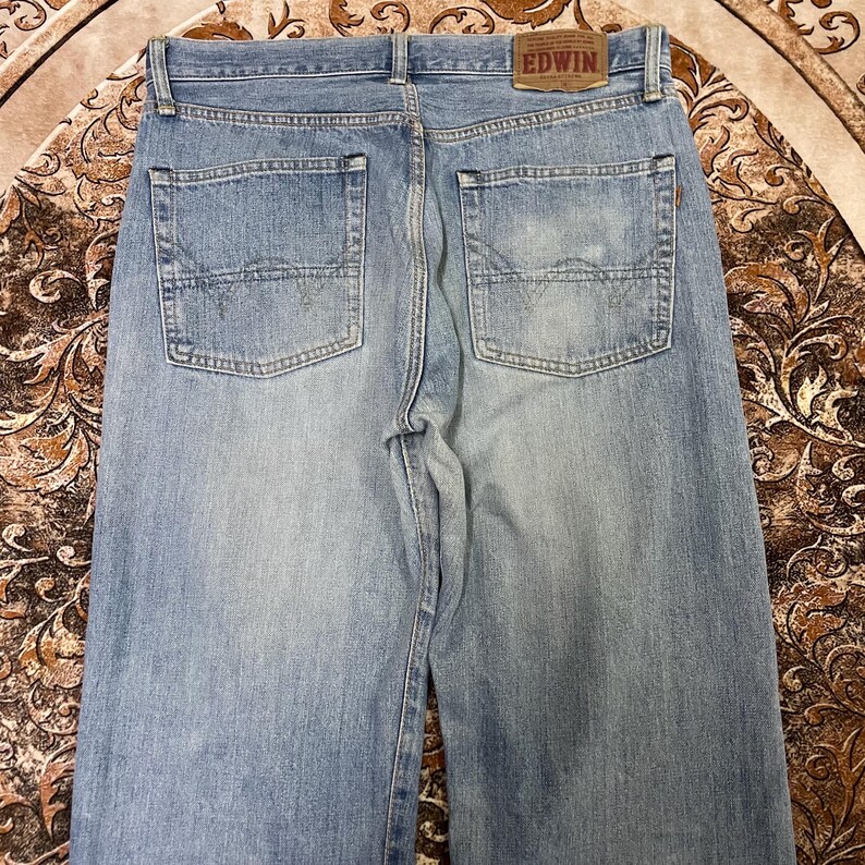 May include: Light blue denim jeans with two back pockets and a brown leather patch with the brand name "EDWIN". The jeans have a classic five-pocket design and a straight leg cut. The denim has a faded wash.