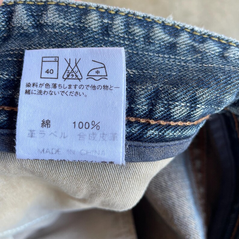 May include: Close-up of a white tag with Japanese text and care symbols, attached to blue denim fabric. The tag indicates the fabric is 100% cotton and provides washing instructions. The tag reads "MADE IN CHINA".