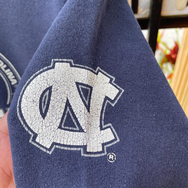 May include: Close-up of a navy blue sweatshirt sleeve featuring a distressed white logo. The logo is a stylized letter "N" and "C" with a registered trademark symbol. The fabric has a slightly worn appearance.