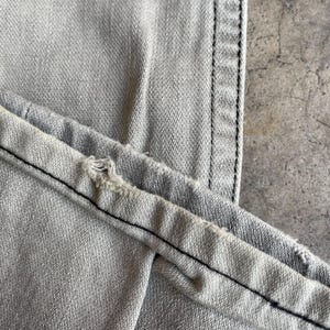 K&ouml;nnte beinhalten: Nahaufnahme einer hellgrauen Jeans aus Denim mit schwarzen N&auml;hten. Die Jeans weist Gebrauchsspuren auf, mit ausgefranstem Rand und sichtbaren Nahtdetails. Der Stoff hat ein strukturiertes Aussehen.