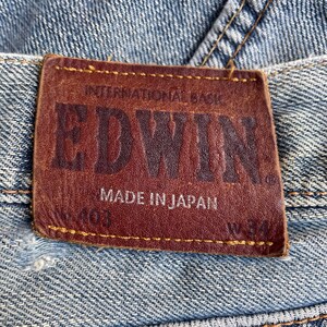 May include: Close-up of a brown leather label on blue denim jeans. The label reads "EDWIN" in embossed letters, with "INTERNATIONAL BASIC" above and "MADE IN JAPAN" below. The jeans have a worn appearance.