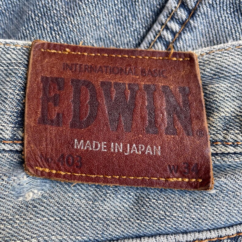 May include: Close-up of a brown leather label on blue denim jeans. The label reads "EDWIN" in embossed letters, with "INTERNATIONAL BASIC" above and "MADE IN JAPAN" below. The jeans have a worn appearance.