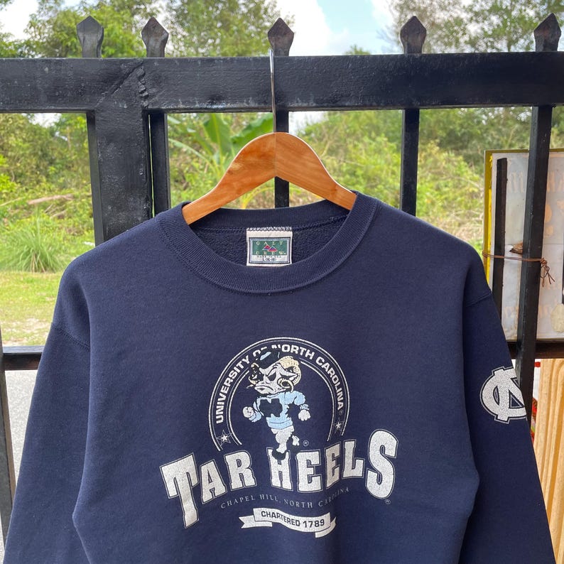 May include: Navy blue sweatshirt with the words "Tar Heels" in white block letters. The sweatshirt features a graphic of a cartoon ram and the words "University of North Carolina" in a circular design. A logo is on the right sleeve.