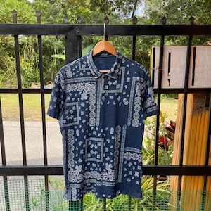 以下が含まれることがあります： 白のペイズリー柄のネイビーブルーのポロシャツ。 シャツは半袖と襟付きです。 シャツは木製のハンガーに吊るされています。