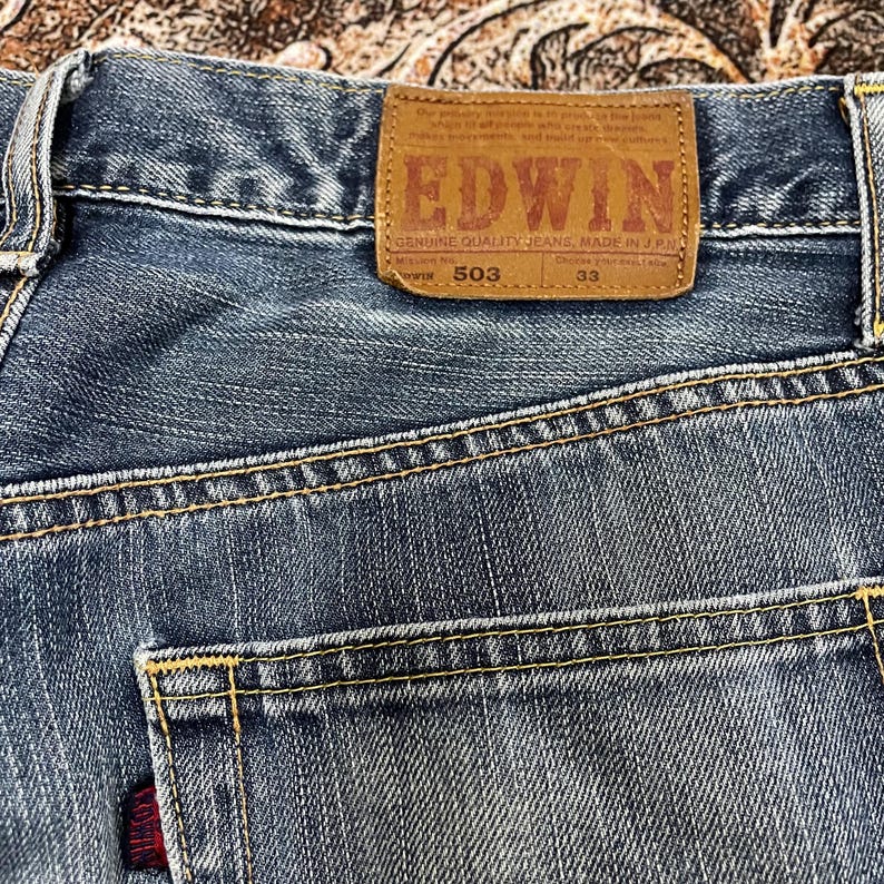 May include: Blue denim jeans featuring a tan leather patch on the back waistband with the text EDWIN, GENUINE QUALITY JEANS, MADE IN J.P.N, 503, and 33. The jeans have yellow contrast stitching, a back pocket, and a small red brand tag.