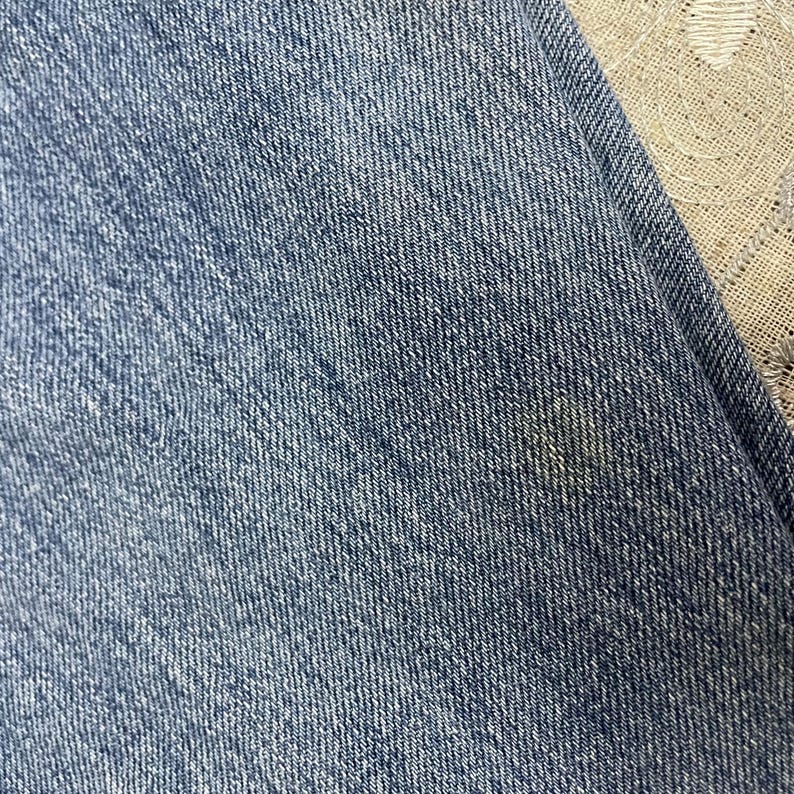 May include: A close-up view of blue denim fabric featuring a classic twill weave pattern. The material displays a medium blue wash with subtle variations in color intensity across the surface, showing the characteristic texture of cotton denim jeans.